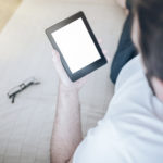Man Relaxing On Couch Using E-book Reader Or Small Tablet Comput