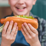 Close-up Partial View Of Smiling Little Boy Holding Hot Dog